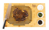 Bamboo carving board with grilled steak, chef knife, and bowls of salt, herbs, and peppercorns