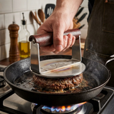 Hand pressing a stainless steel grill press on a burger patty in a cast iron skillet on stove
