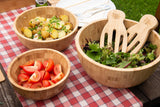 Three bamboo wooden salad bowls with matching salad servers on a picnic table, filled with fresh greens, tomatoes, and potato salad.