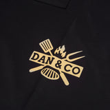Black kitchen apron with yellow grill logo, spatula, fork, and flame graphic