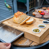 Bamboo bread cutting board with crumb tray, sliced bread, olives, and dipping oil outdoors