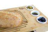 Bamboo bread board with loaf, dips, green olives and black olives in ceramic bowls