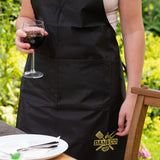 Person wearing black kitchen apron outdoors holding wine glass, table setting visible