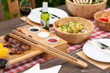 Outdoor table with bamboo serving trays, bowls, grilled food and picnic tableware