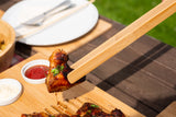 Bamboo tongs grip grilled chicken wing on bamboo carving board with dip bowls outdoors